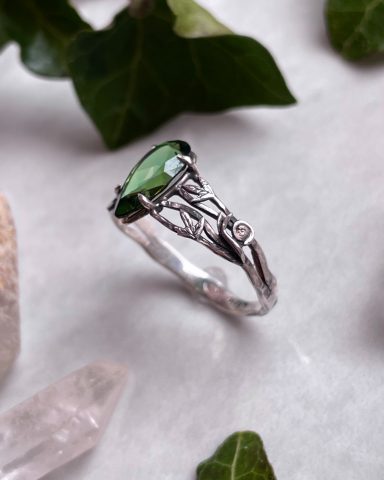 Silberner Ring mit grünem Edelstein, umgeben von grünen Blättern und Kristallen.