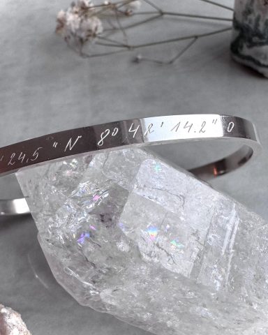Silberner Armband mit geprägten Koordinaten und klaren Kristallen im Hintergrund.